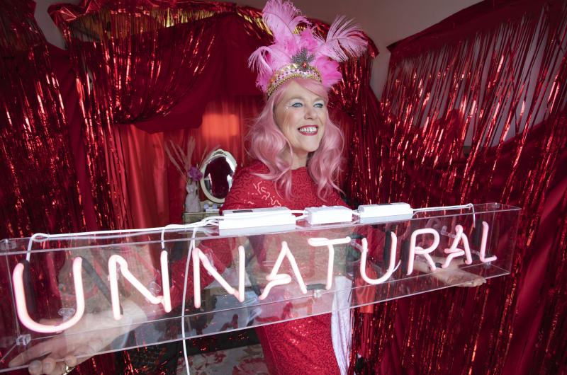 An image of a woman standing in front of a glitter fringe curtain decorated beauty salon, with a neon sign saying 'Unnatural"