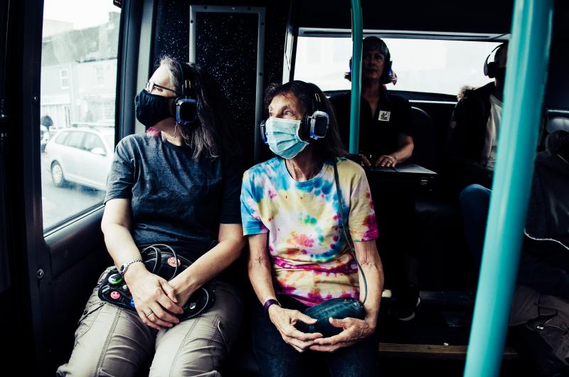 An image of two people sitting on a bus looking out of the window, with a headset on
