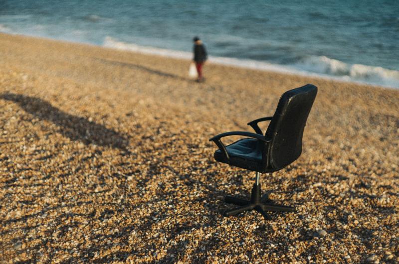 An image of a chair on a pebble beach with the sea in the background and a person walking past