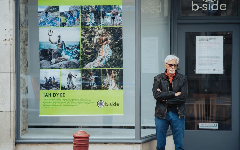 A man in sunglasses and leather jacket stands outside a large window with a colourful photographic poster