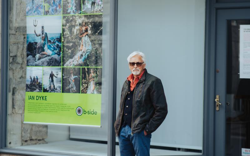 A man with grey hair and sunglasses standing beside a large colourful poster